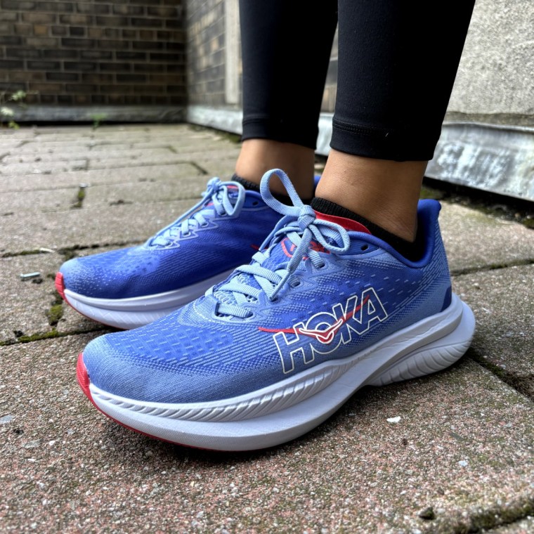 A woman standing on a brick floor while modeling a blue pair of the Hoka Gaviota 5.