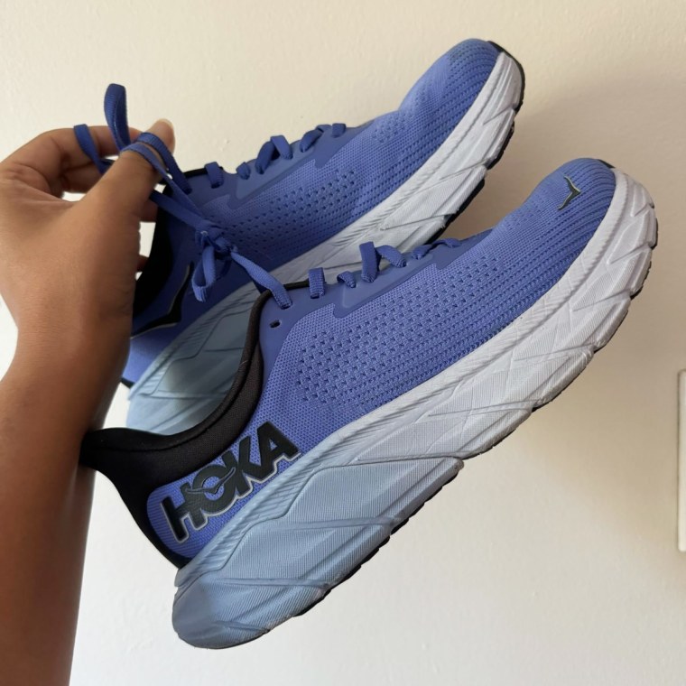 A woman’s hand holding up a blue pair of Hoka Arahi 7s against a white blank wall.