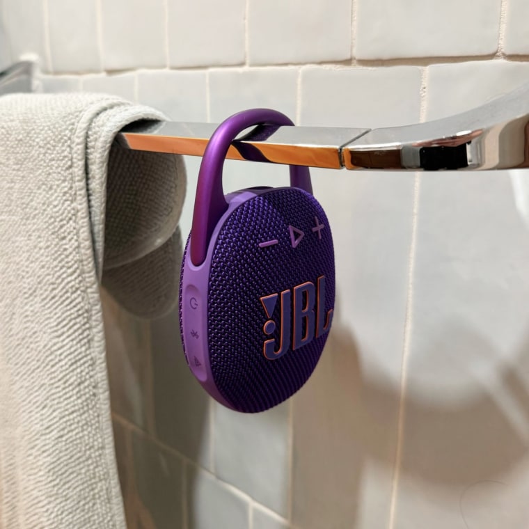 Purple oval shaped JBL speaker clipped on to towel rod in a bathroom.