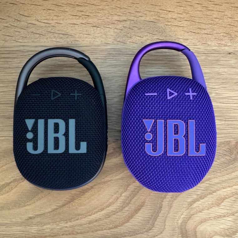 Purple oval-shaped JBL speaker with carabiner clip on the right and slightly smaller black oval-shaped JBL speaker with carabiner clip on the left.