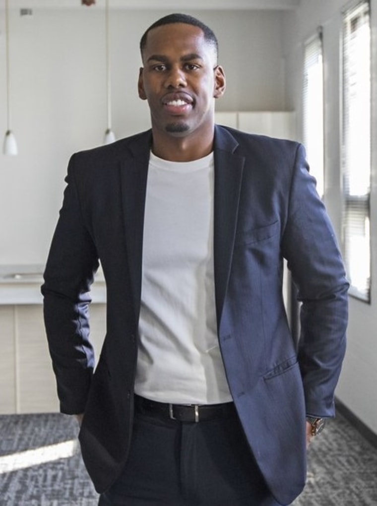 Black man wearing white t-shirt and navy jacket standing in white office