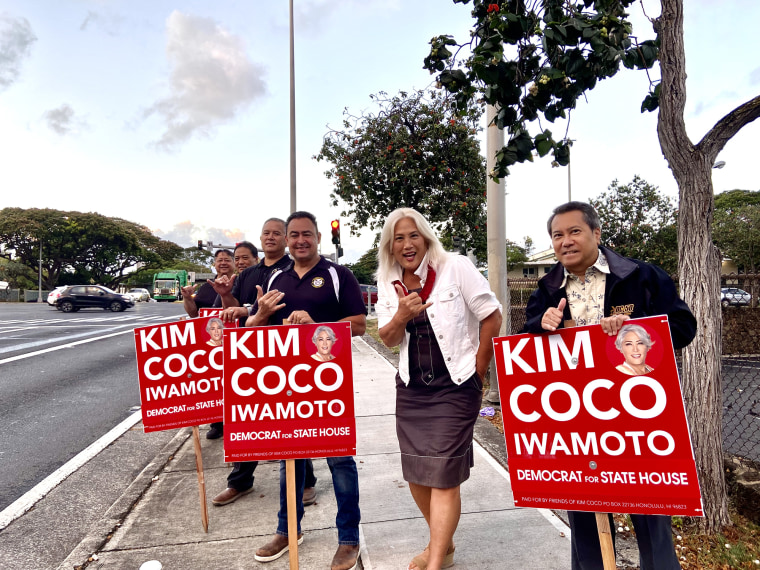 Photo of subject Kim Coco Iwamoto standing on a sidewalk with 5 other people who each old red campaign signs with her name 