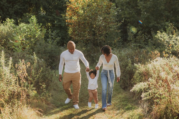 A dad, son, and mother hold hands and walk down a grassy path lined with bushes